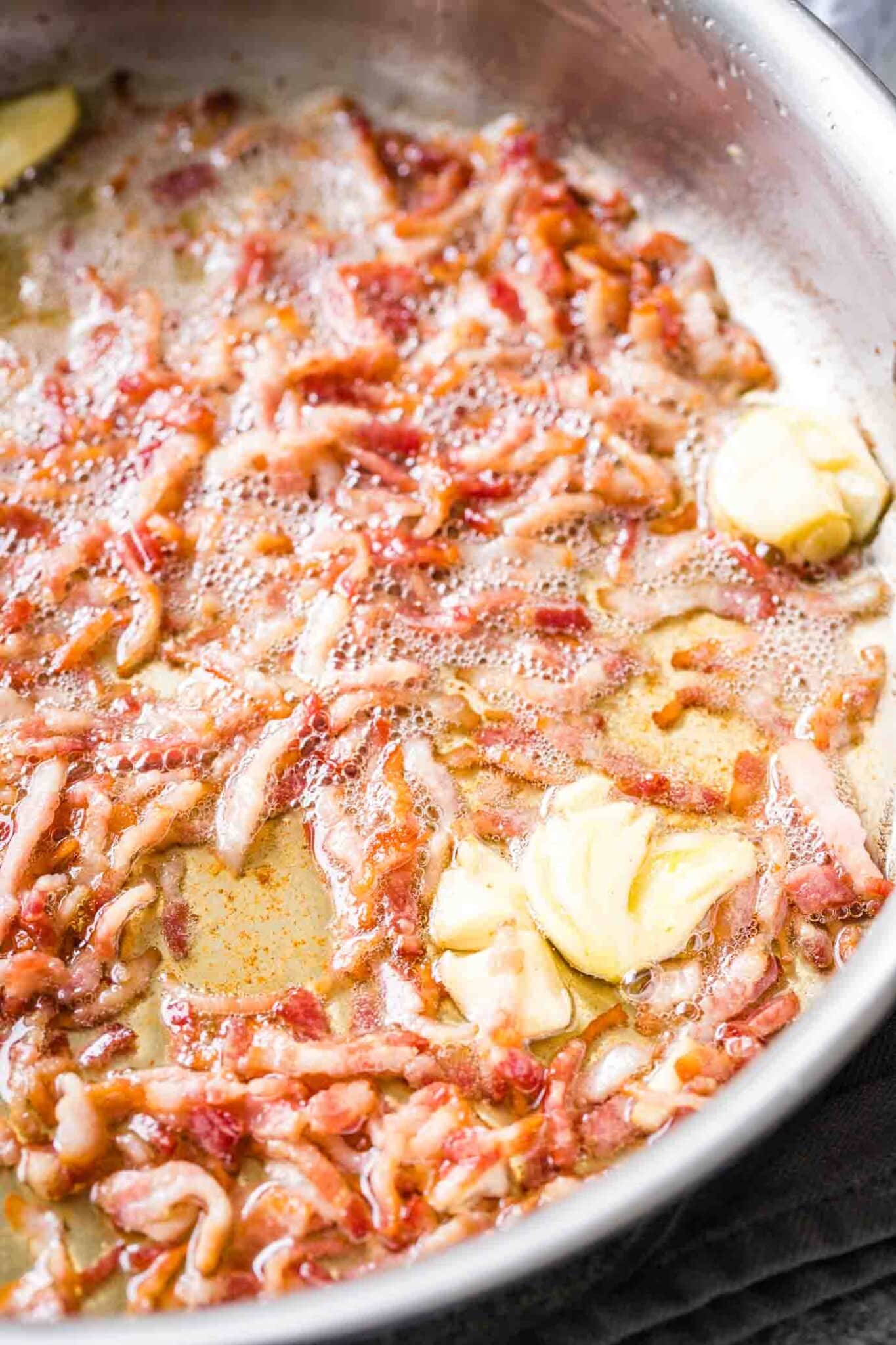Bacon, coupé en petits morceaux, en train de frémir dans une poêle en acier inoxydable, avec des gousses d'ail écrasées et pelées.