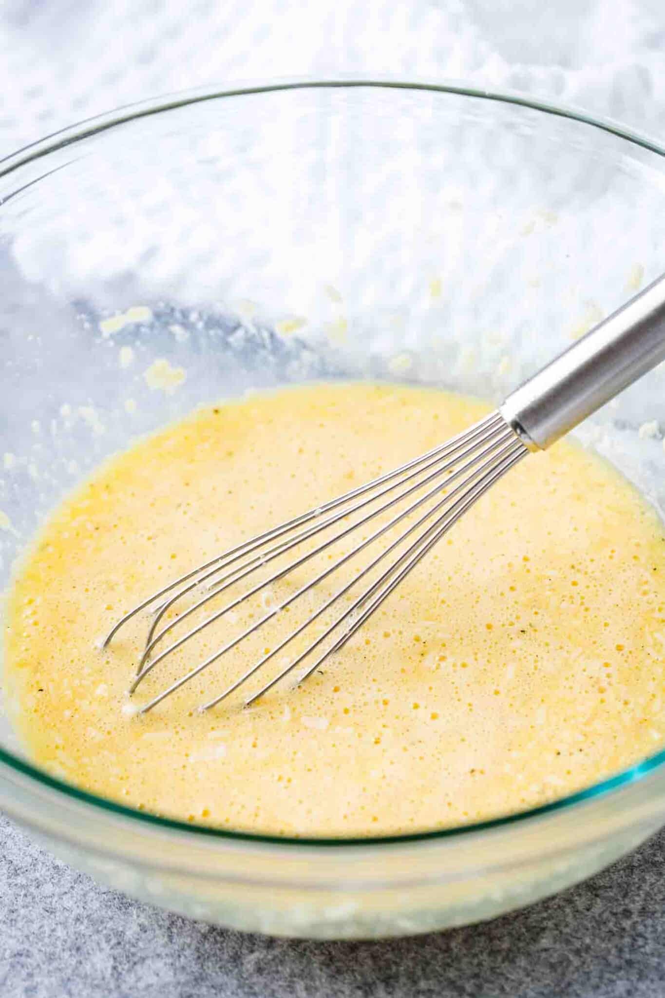 Un bol en verre avec un fouet en acier inoxydable contenant un mélange d'œufs et de parmesan râpé.