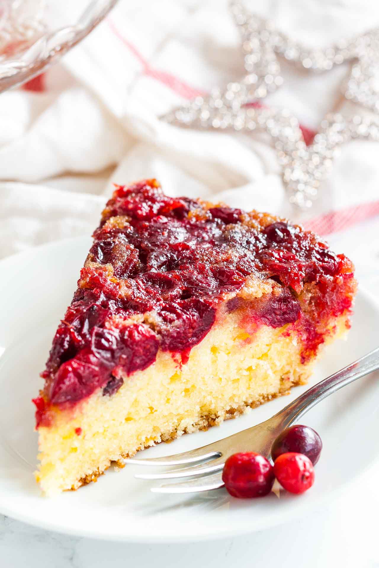 Une tranche de gâteau renversé aux canneberges sur une assiette blanche avec une fourchette, garnie de canneberges et d'étoiles de Noël en arrière-plan