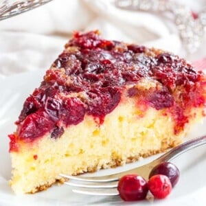 Une tranche de gâteau renversé aux canneberges sur une assiette blanche avec une fourchette, garnie de canneberges et d'étoiles de Noël en arrière-plan