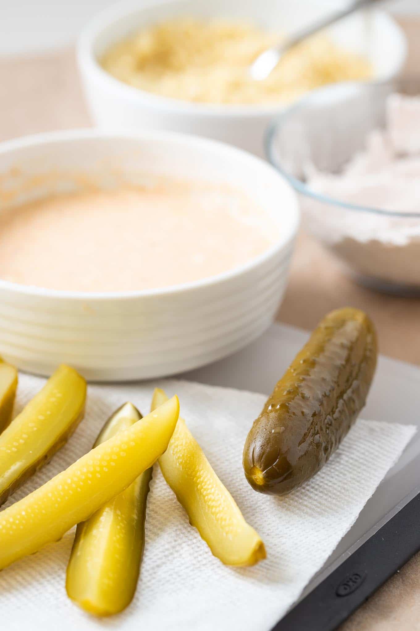 Les cornichons à la friteuse à air sont super croustillants et constituent un excellent en-cas pour le jour de match ou une fête ! Les cornichons frits à la friteuse à air sont plus sains car ils sont faits avec beaucoup moins d'huile et ont une couche de chapelure panko qui les rend encore meilleurs que l'original frit. #RecettesFriteuseÀAir #CornichonsFrits Quartiers de cornichons à l'aneth, avec un cornichon entier sur une planche à découper avec un torchon. Il y a un bol blanc avec de la pâte et un bol en verre avec de la farine en arrière-plan.