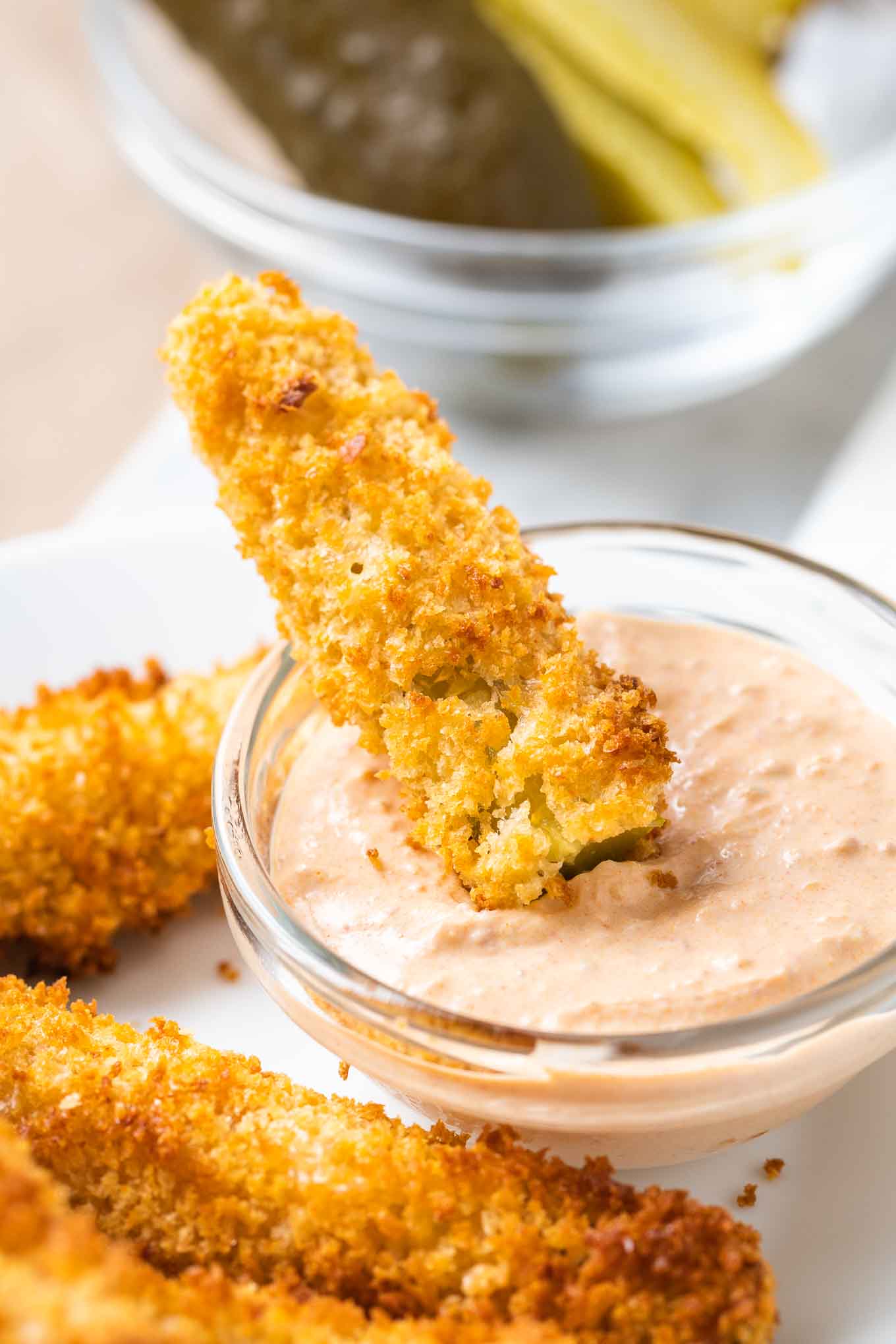 Les cornichons à la friteuse à air sont super croustillants et constituent un excellent en-cas pour le jour de match ou une fête ! Les cornichons frits à la friteuse à air sont plus sains car ils sont faits avec beaucoup moins d'huile et ont une couche de chapelure panko qui les rend encore meilleurs que l'original frit. #RecettesFriteuseÀAir #CornichonsFrits Un cornichon frit est plongé dans une sauce dans un petit bol en verre, avec d'autres cornichons frits à côté sur une assiette blanche.