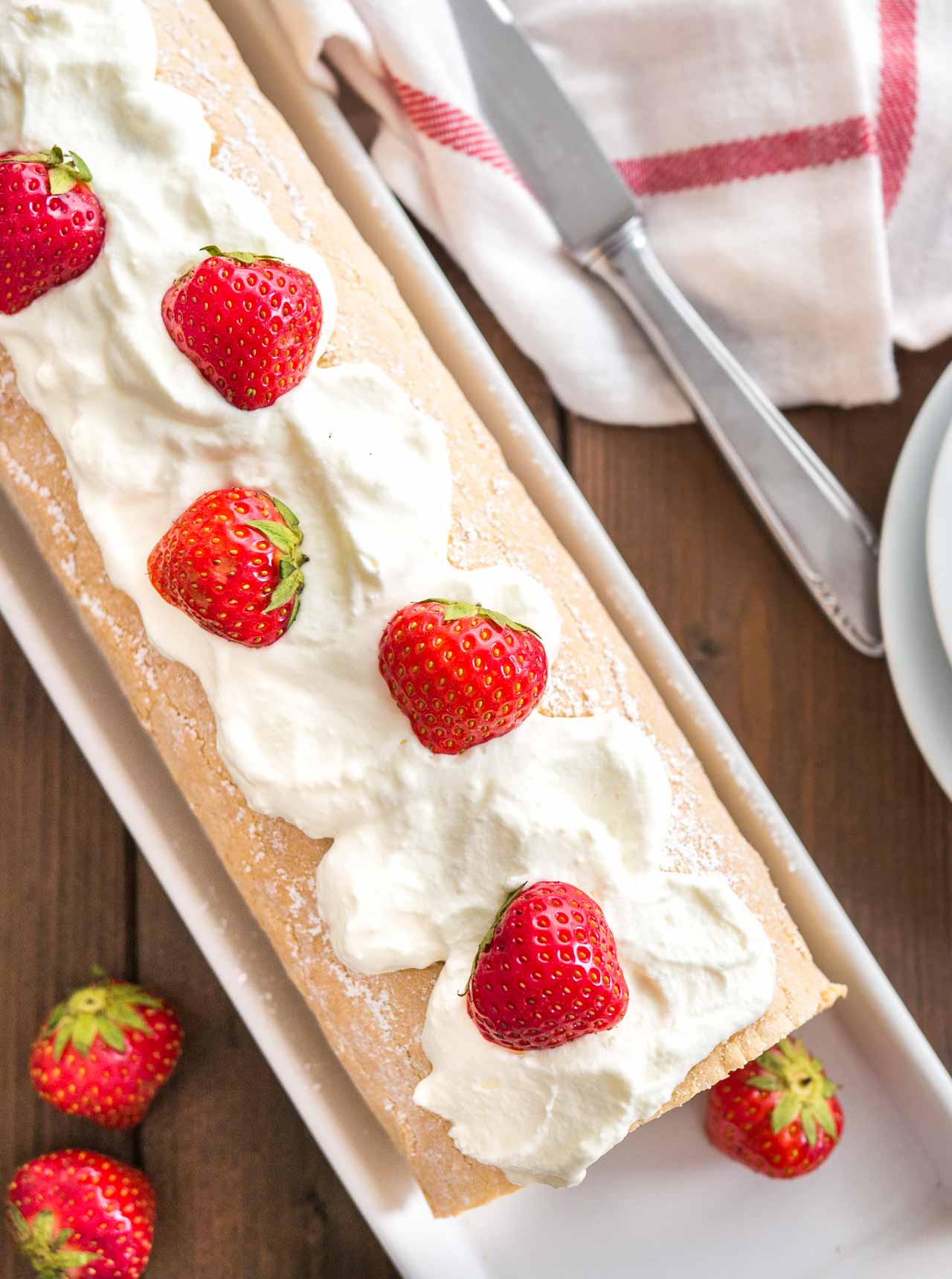 Vue de dessus d'un gâteau roulé aux fraises surmonté de crème fouettée et de fraises, sur un plat de service rectangulaire. Des assiettes, un couteau et un torchon blanc et rouge sont à côté.