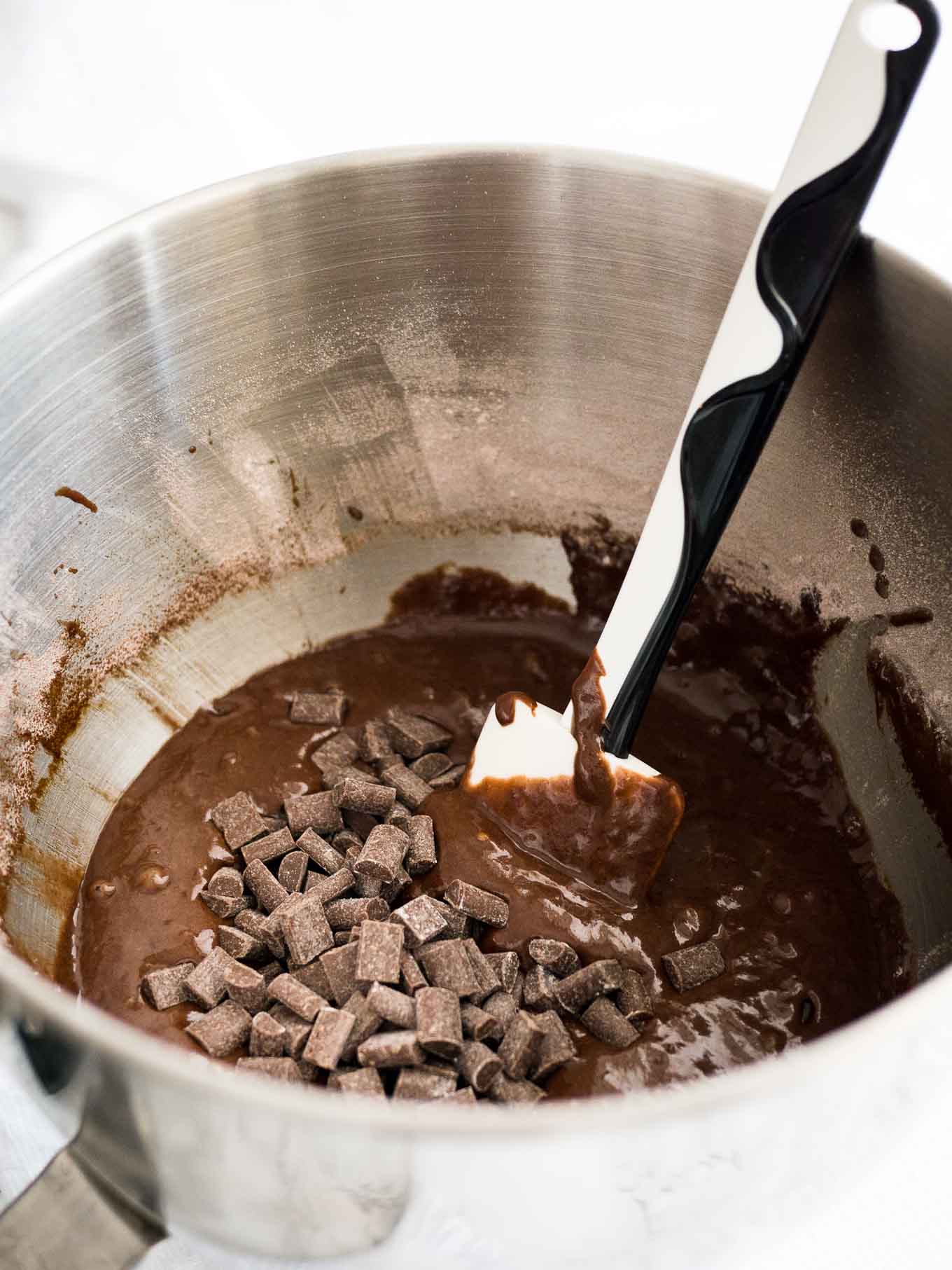 Un bol en acier inoxydable avec une pâte chocolatée et des pépites de chocolat, avec une spatule noir et blanc dedans.