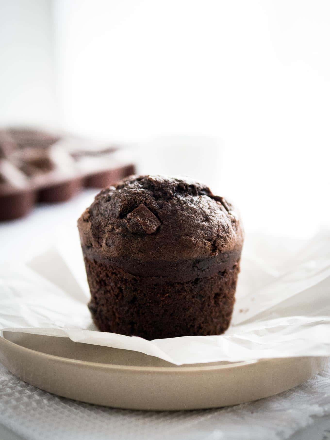 Gros plan sur un muffin chocolat-banane sur une assiette grise avec du papier sulfurisé, avec d'autres muffins en arrière-plan.
