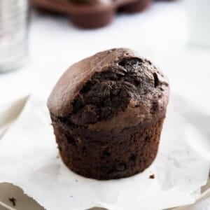 Un muffin chocolat-banane sur une assiette grise avec du papier sulfurisé à côté d'une cuillère en bronze et un verre avec d'autres muffins en arrière-plan.