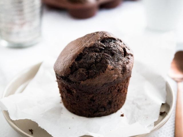 Un muffin chocolat-banane sur une assiette grise avec du papier sulfurisé à côté d'une cuillère en bronze et un verre avec d'autres muffins en arrière-plan.