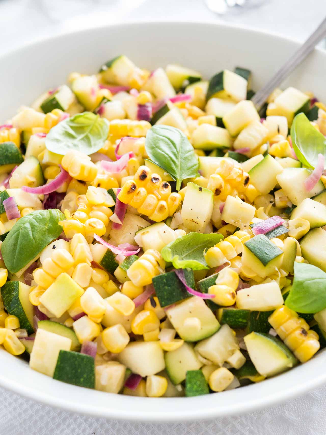 Gros plan de salade de courgettes et maïs rôties avec des oignons dans un bol blanc, garnie de basilic, avec une cuillère dedans.