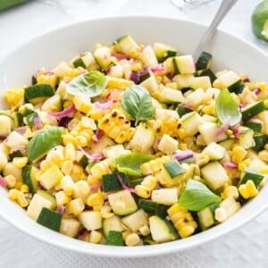 Salade de courgettes et maïs rôties avec des oignons dans un bol blanc, garnie de basilic, avec une cuillère dedans