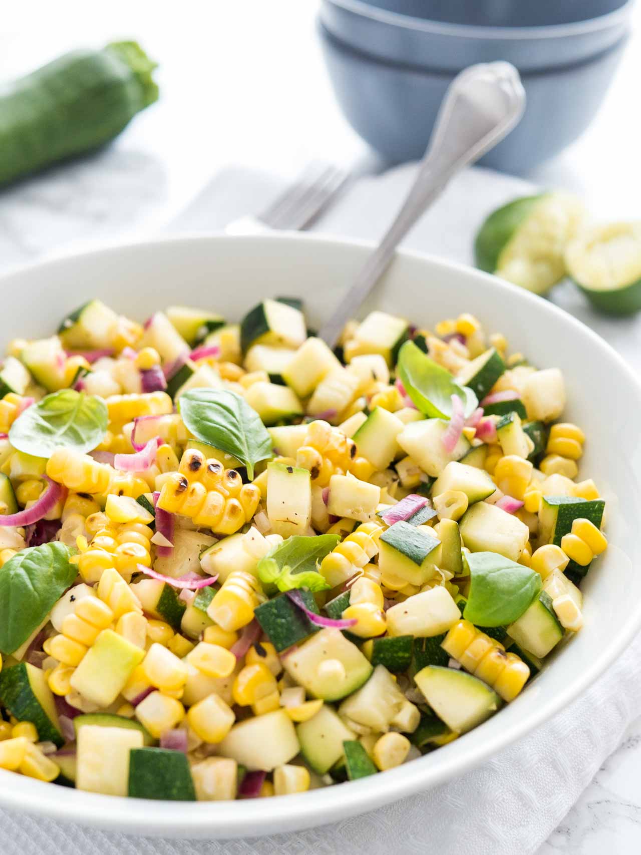 Salade de courgettes et maïs rôties avec des oignons dans un bol blanc, garnie de basilic, avec une cuillère dedans sur une table blanche avec une fourchette, des courgettes et des citrons verts pressés en arrière-plan.