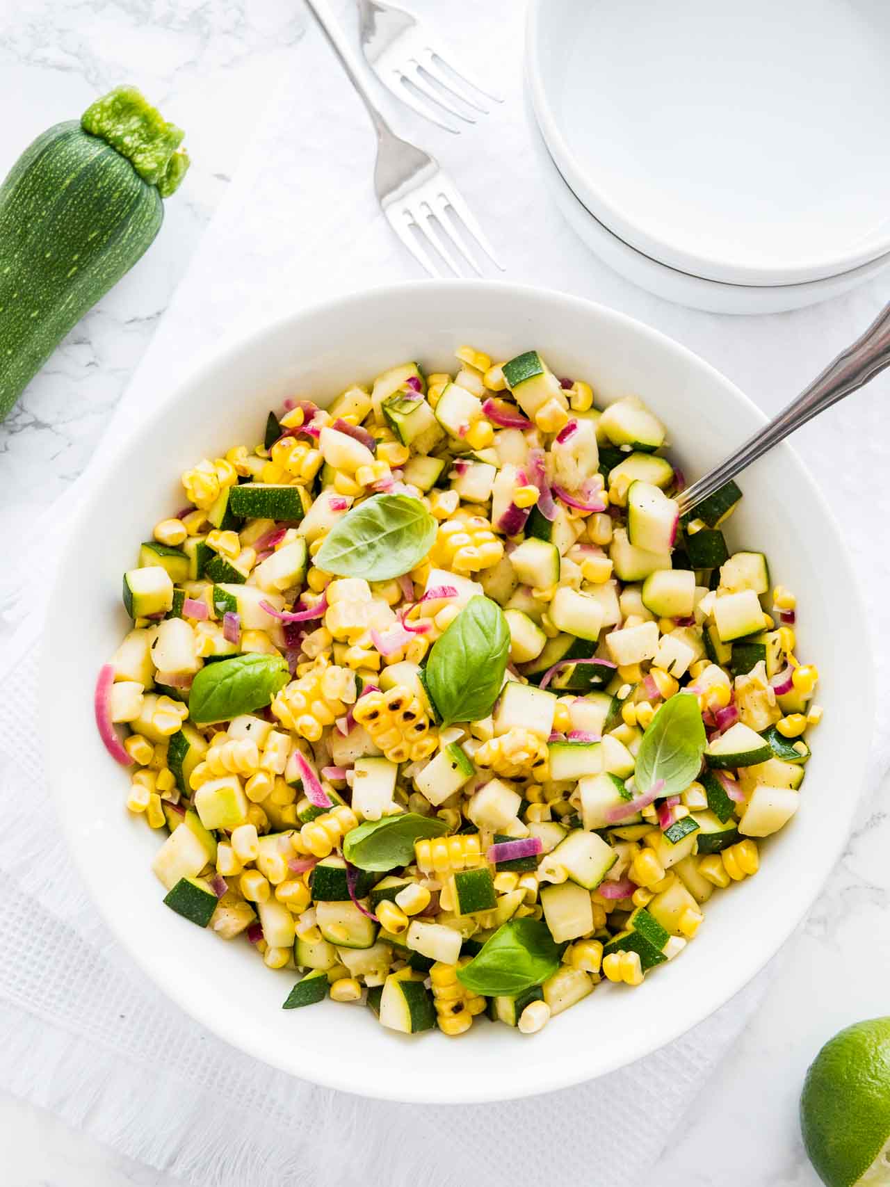 Salade de courgettes et maïs dans un bol blanc, garnie de basilic, avec une cuillère dedans sur une table blanche avec une fourchette et une courgette à côté.