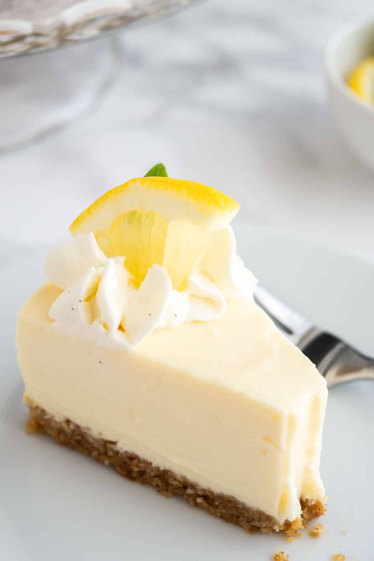 Gros plan d'une part de tarte à la crème au citron sur une assiette blanche avec une fourchette sur une surface en marbre.