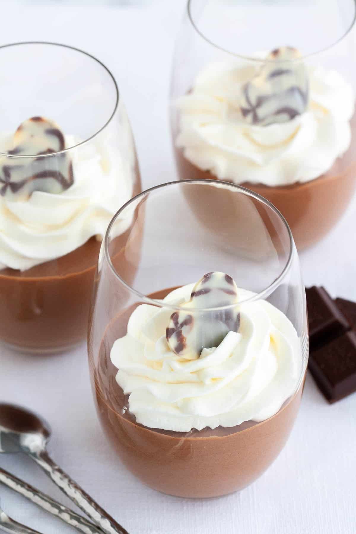 Cette recette de mousse au chocolat est super simple à réaliser maison et délicieusement légère et aérée. Une recette de mousse sans œuf qui est si aérienne et chocolatée et peut être appréciée par tout le monde. Assez élégante pour une fête, mais suffisamment facile à préparer chaque fois que vous avez une envie de chocolat ! Trois verres de mousse au chocolat, surmontés de crème fouettée à côté de morceaux de chocolat et d'une cuillère.