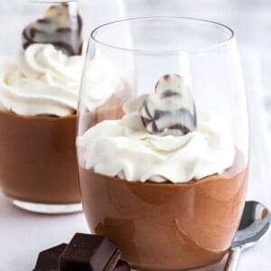 Mousse au chocolat dans un verre, surmontée de crème fouettée à côté de morceaux de chocolat et d'une cuillère.