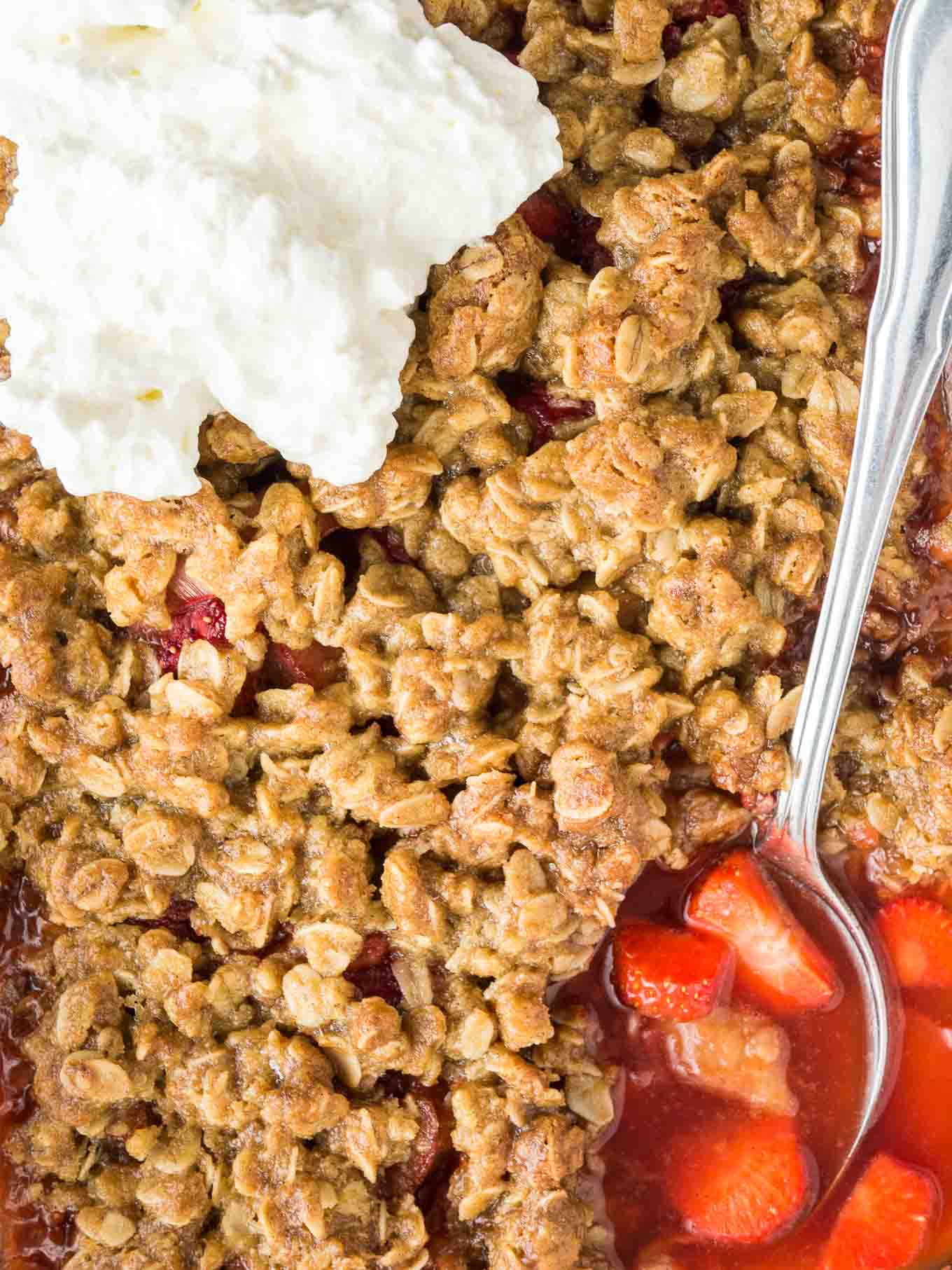 Crumble aux fraises et à la rhubarbe surmonté de crème, avec une cuillère creusant dans la couche inférieure de rhubarbe et de fraises.