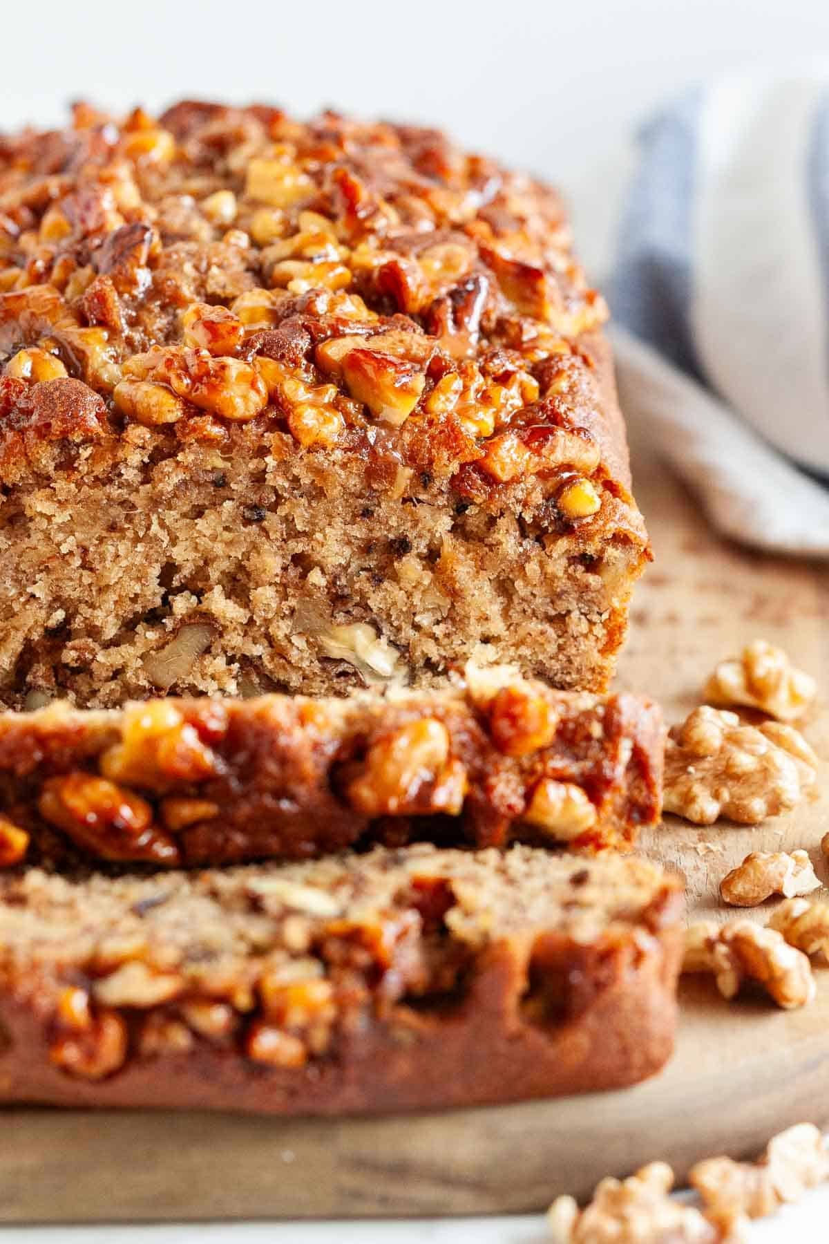 Ce pain à la banane est très moelleux et délicieux ! Surmonté de noix caramélisées et chargé de noix et de bananes écrasées, ma recette de pain à la banane est un favori de la famille qui est parfait pour le petit-déjeuner ou comme collation. Une façon idéale d'utiliser des bananes trop mûres ! #painbanane #painbananeauxnoix #recettesbananes #bananes #pain #recettespetitdéjeuner Un pain à la banane tranché sur une planche en bois.