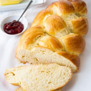 Un Hefezopf, avec une tranche coupée et posée devant sur un torchon blanc à côté d'un petit bol blanc de confiture de fraises avec une cuillère.