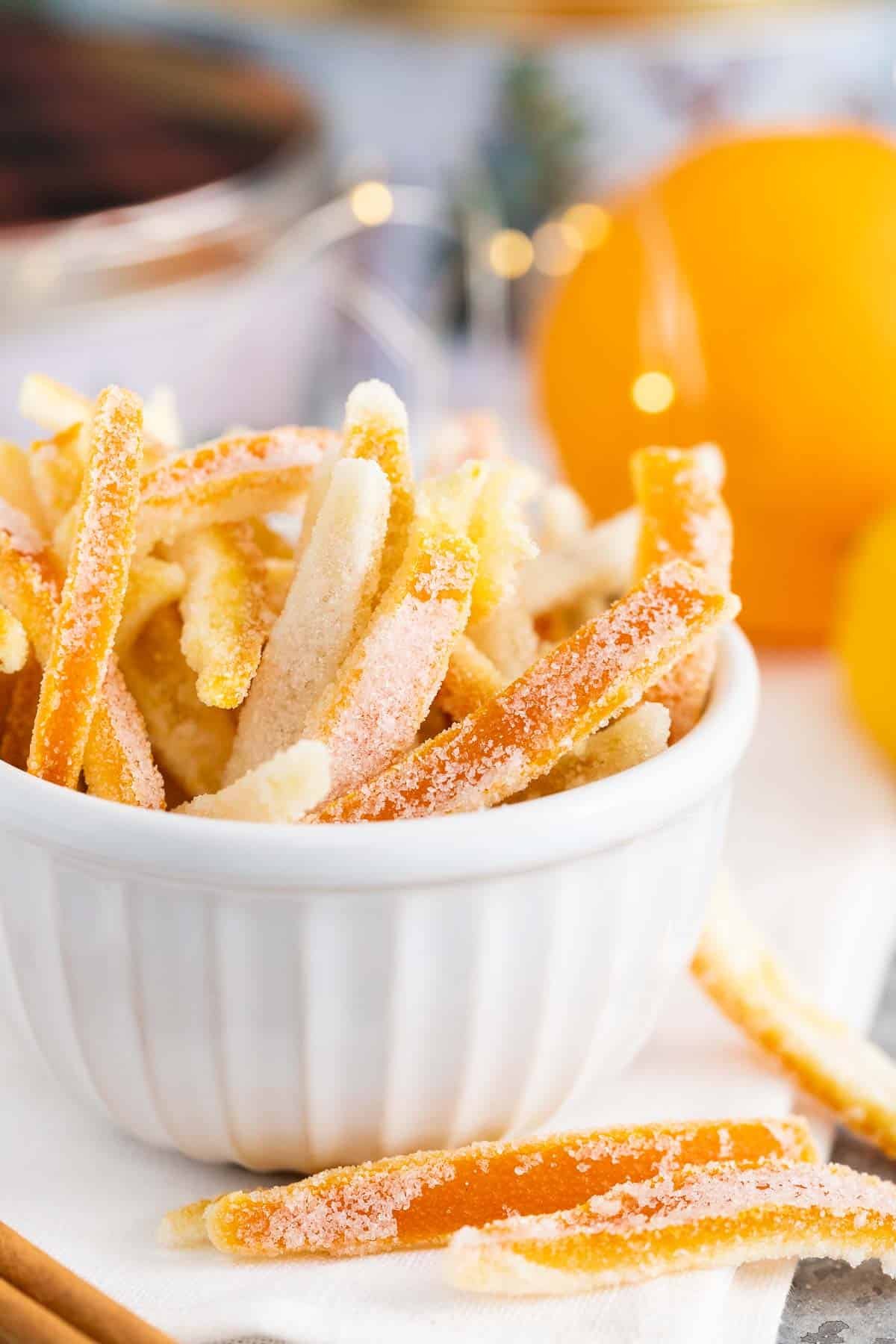 Écorces confites dans un petit bol blanc avec des oranges en arrière-plan.