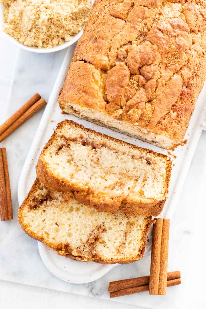 Vue de dessus du pain au cannelle sur un plat de service rectangulaire blanc, avec 2 tranches coupées et posées, agrémenté de bâtons de cannelle.