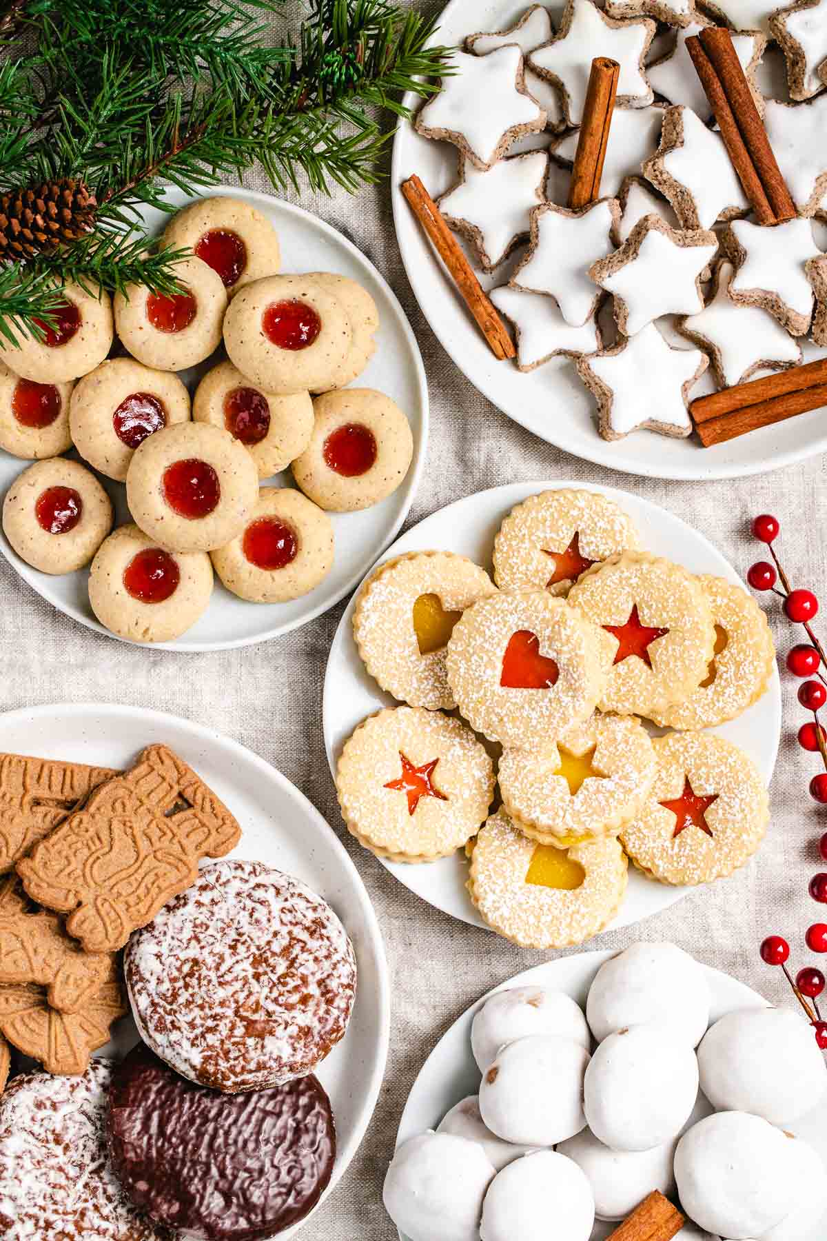 Une collection de biscuits de Noël allemands sur diverses assiettes garnies de branches de pin et de baies rouges. Il y a des étoiles à la cannelle, garnies de bâtons de cannelle, des biscuits à empreintes avec de la confiture de fraise, des biscuits Linzer avec de la confiture de fraise et du lemon curd, des biscuits en pain d'épice et des speculoos, et des Pfeffernüsse.