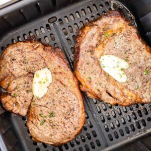 Deux steaks cuits avec un peu de beurre à l'ail sur le dessus dans un panier noir pour air fryer.