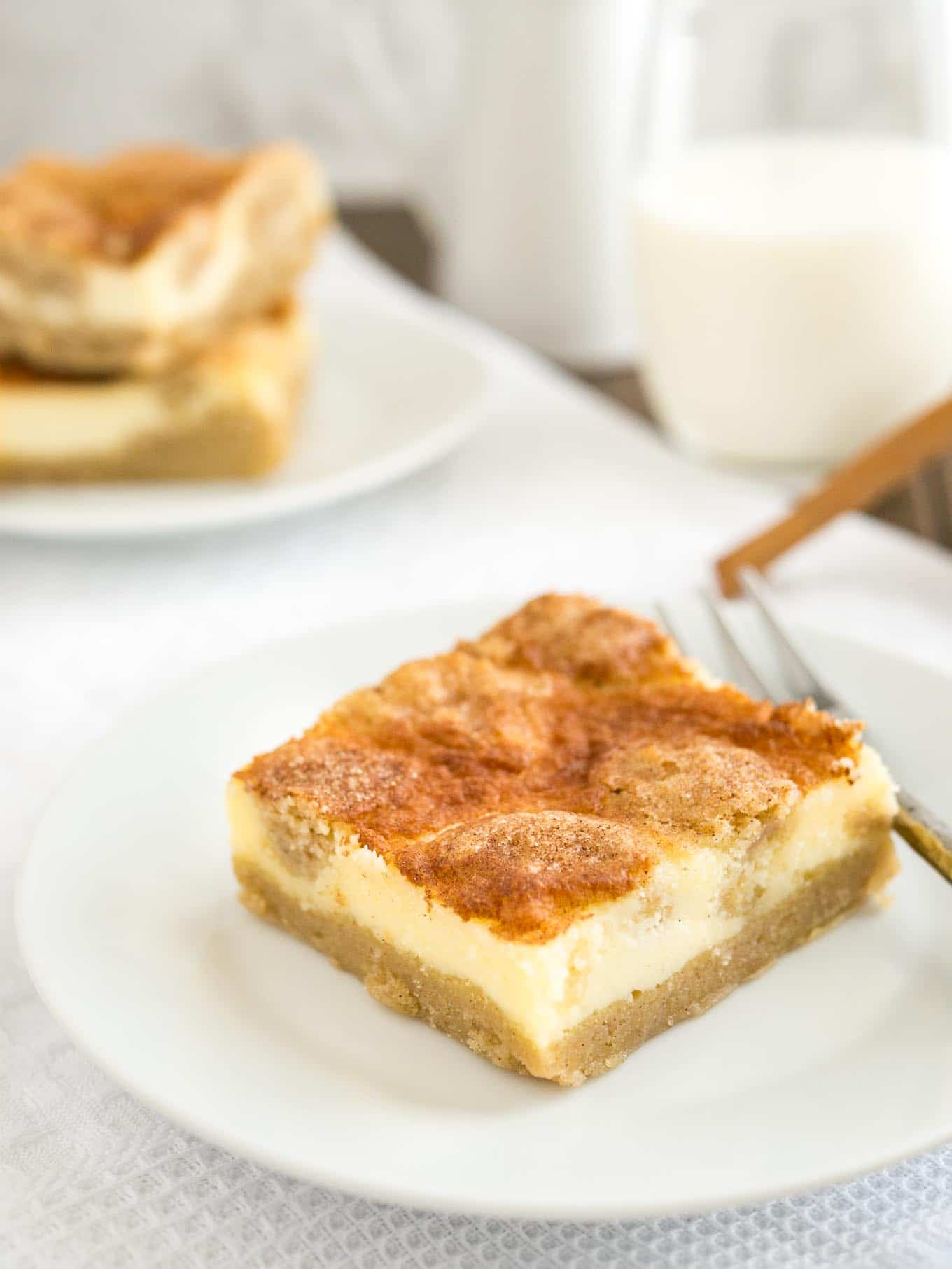 Une assiette blanche avec une barre de cheesecake snickerdoodle et une fourchette dessus sur une nappe blanche. Il y a un verre de lait et une autre assiette avec deux barres de cheesecake en arrière-plan.