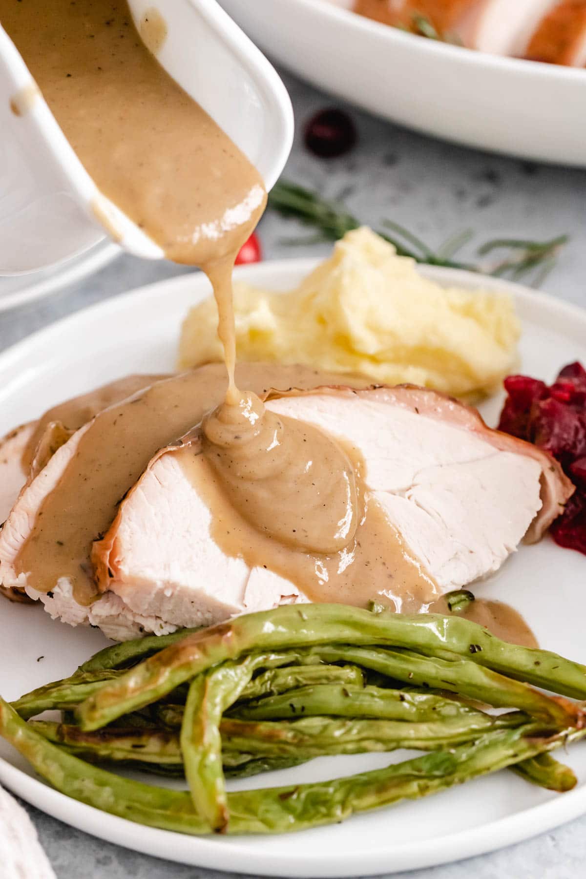De la sauce versée sur une tranche de poitrine de dinde à côté de haricots verts, de purée de pommes de terre et de sauce aux canneberges sur une assiette blanche.