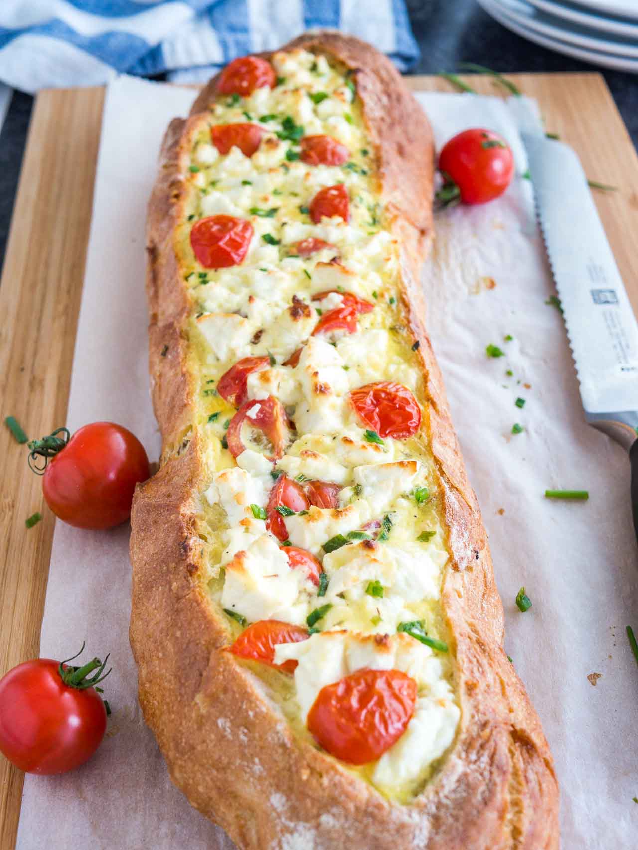 Pain français farci à la tomate et à la feta garni de tomates et de ciboulette sur une planche à découper en bambou recouverte de papier sulfurisé. Un couteau à pain est posé à côté.