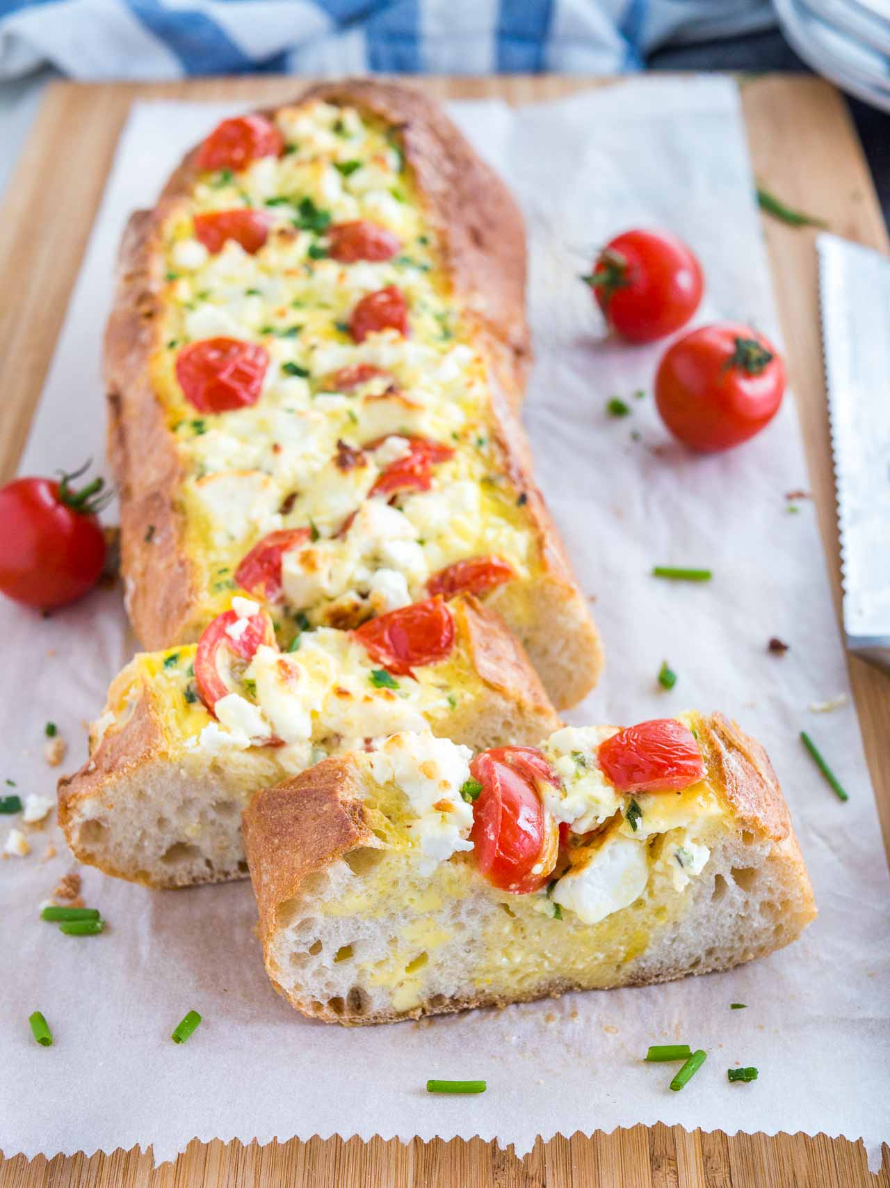 Pain français farci à la tomate et à la feta garni de tomates et de ciboulette sur une planche à découper en bambou recouverte de papier sulfurisé. Deux tranches ont été coupées et sont posées devant lui, et un couteau à pain est à côté.