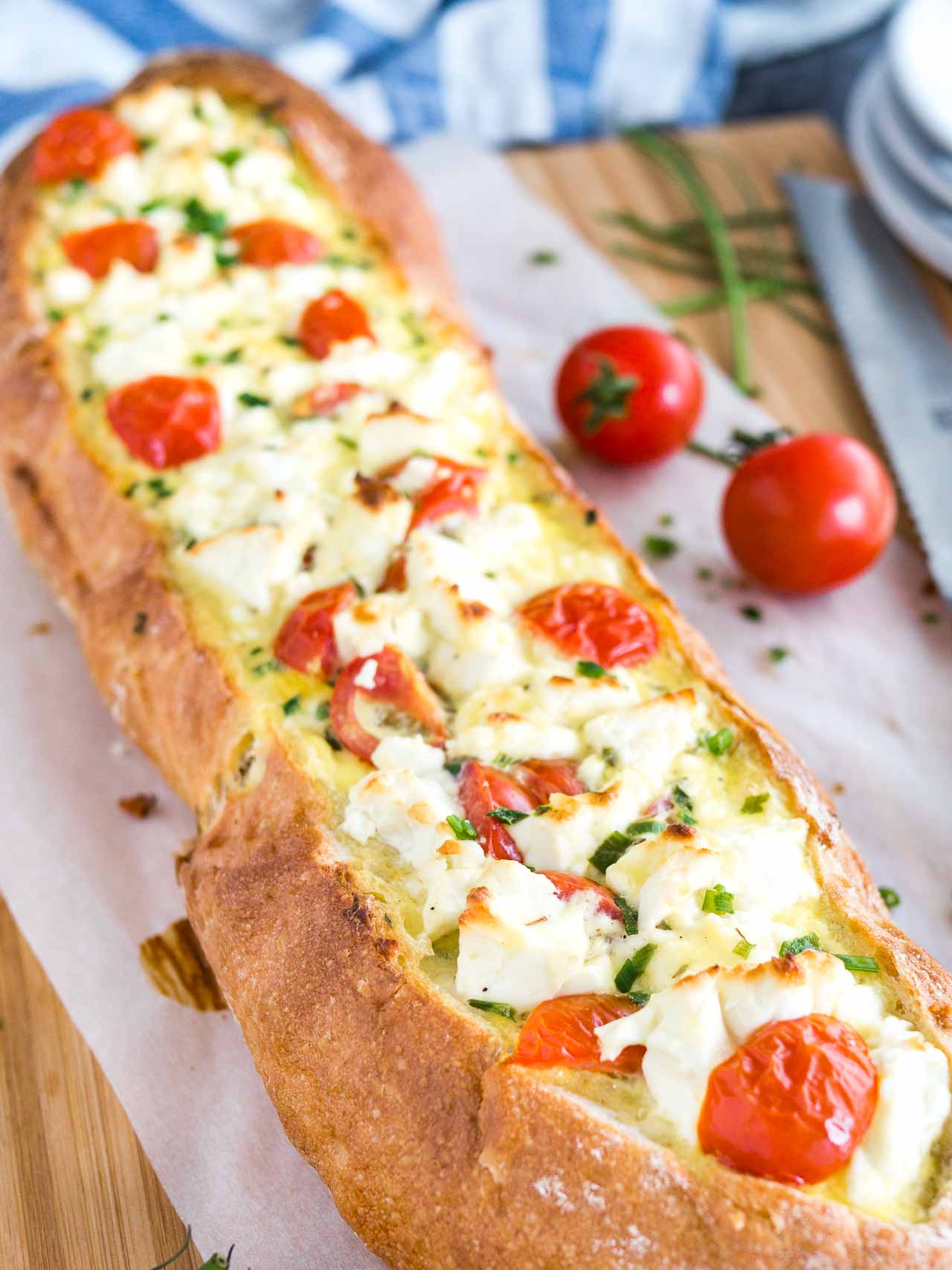 Gros plan sur du pain français farci à la tomate et à la feta garni de tomates et de ciboulette sur une planche à découper en bambou recouverte de papier sulfurisé. Un couteau à pain est posé à côté.