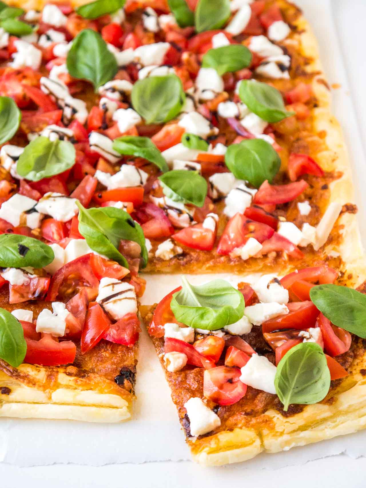 Pizza en pâte feuilletée tomate mozzarella avec tomates, mozzarella, feuilles de basilic et glaçage balsamique, une part a été découpée et est posée devant.