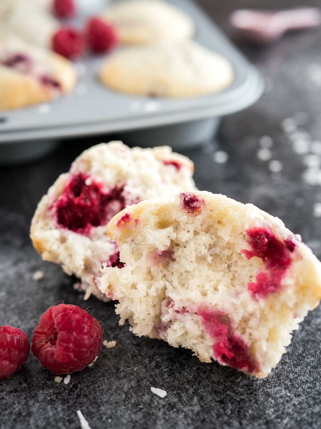 Gros plan d'un muffin à la noix de coco et à la framboise cassé en deux garnis de framboises. Il y a un moule à muffins gris avec d'autres muffins en arrière-plan.