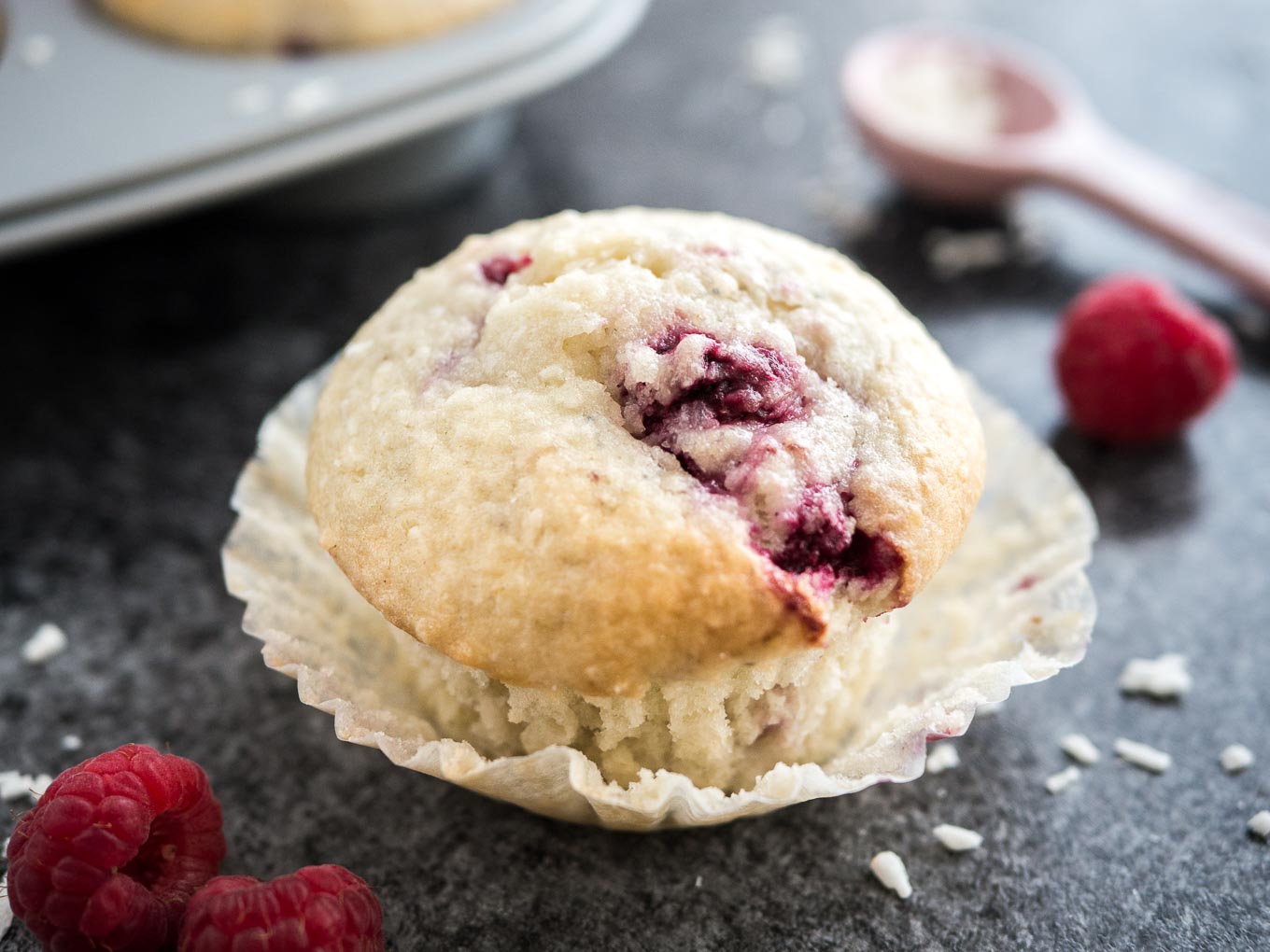 Gros plan d'un muffin à la noix de coco et à la framboise avec son emballage ouvert posé sur une surface sombre. Il y a des framboises, une cuillère à mesurer rose avec des copeaux de noix de coco et un moule à muffins gris avec d'autres muffins à côté.