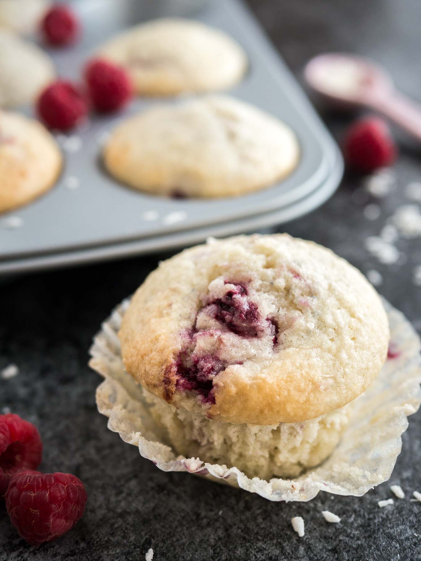 Un muffin à la noix de coco et à la framboise avec son emballage ouvert posé sur une surface sombre. Il y a des framboises, une cuillère à mesurer rose avec des copeaux de noix de coco et un moule à muffins gris avec d'autres muffins à côté.