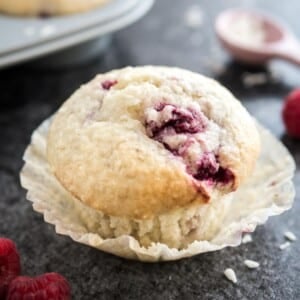 Gros plan d'un muffin à la noix de coco et à la framboise avec son emballage ouvert posé sur une surface sombre. Il y a des framboises, une cuillère à mesurer rose avec des copeaux de noix de coco et un moule à muffins gris avec d'autres muffins à côté.