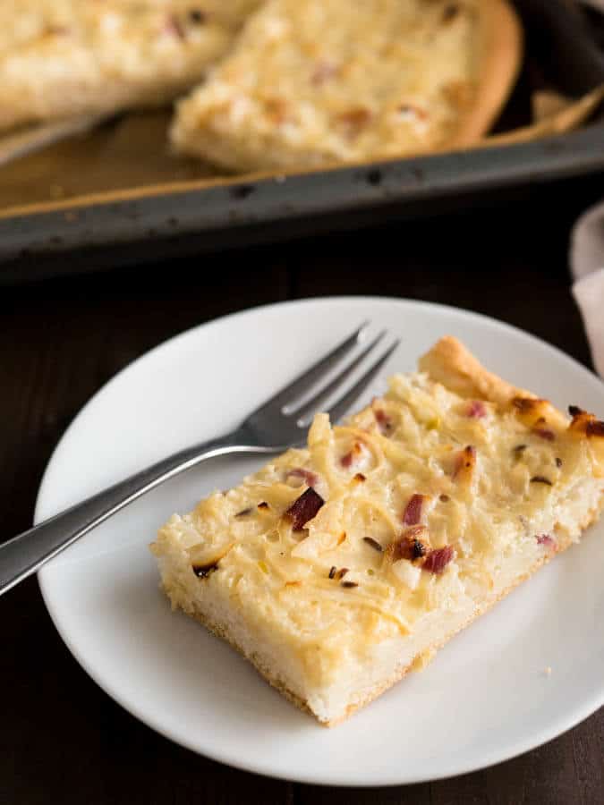 Une assiette blanche avec un morceau de tarte à l'oignon et une fourchette. On voit une plaque de cuisson avec d'autres morceaux de tarte à l'arrière-plan.