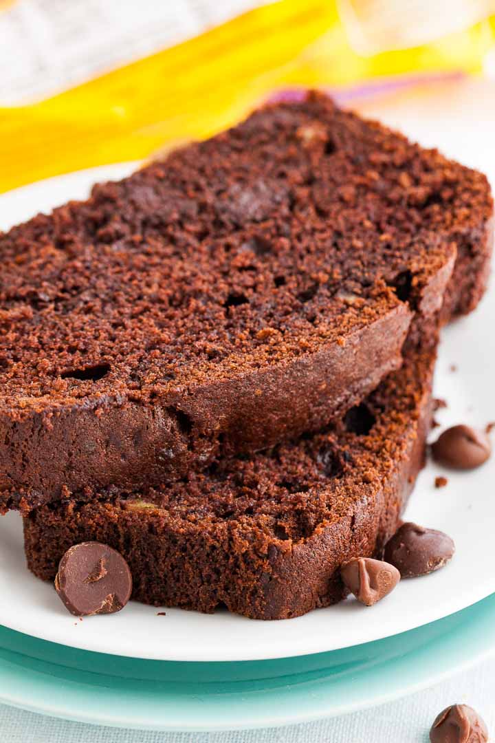 2 tranches de pain à la banane et au chocolat empilées sur une assiette blanche, garnies de pépites de chocolat et avec une banane en arrière-plan.