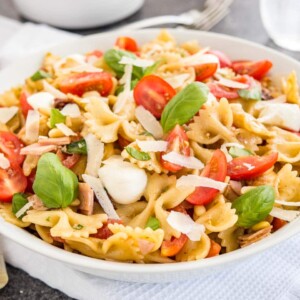 Un bol blanc avec une salade de pâtes avec vinaigrette italienne, avec des tomates, de la mozzarella, des feuilles de basilic et des copeaux de parmesan. Le bol est posé sur un torchon blanc avec des fourchettes, un verre d'eau et des pinces à salade en arrière-plan.