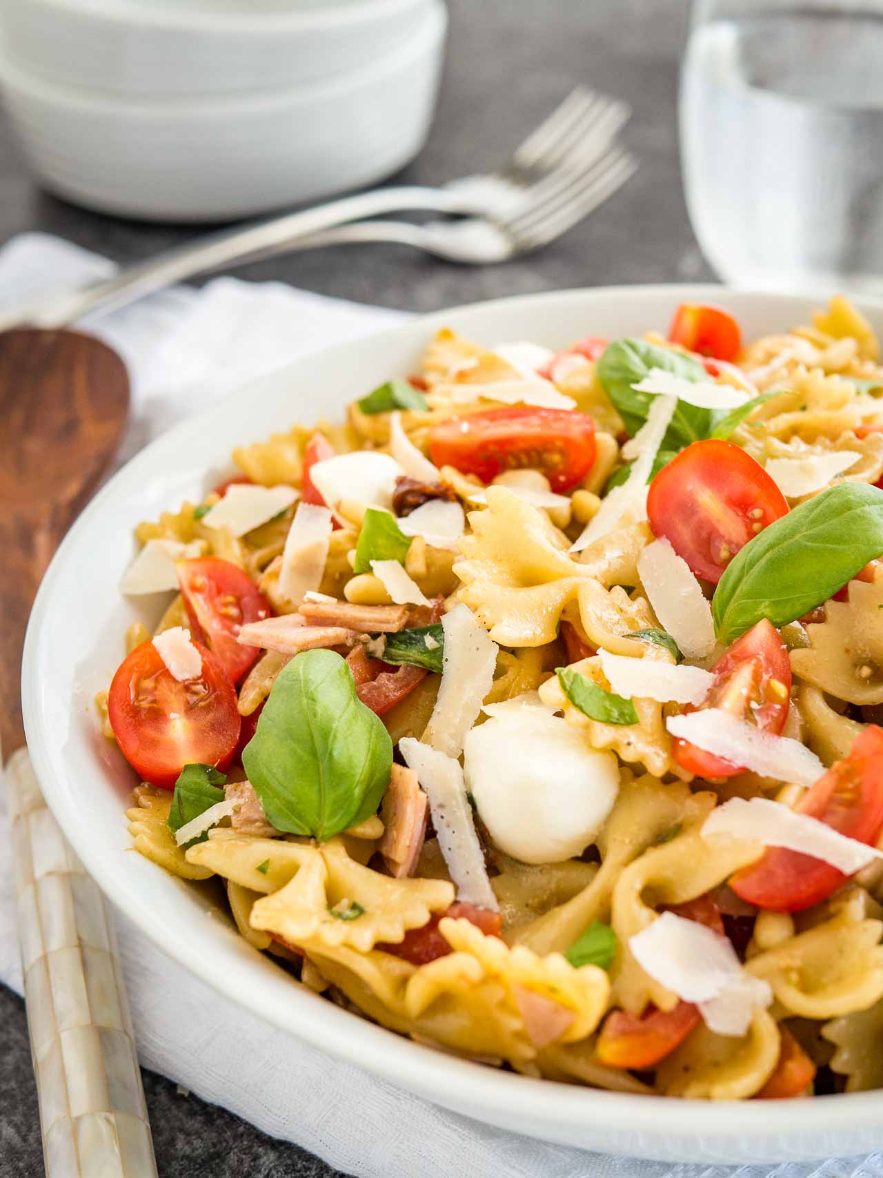 Gros plan sur un bol blanc contenant une salade de pâtes avec vinaigrette italienne, avec des tomates, de la mozzarella, des feuilles de basilic et des copeaux de parmesan. Le bol est posé sur un torchon blanc avec des fourchettes, un verre d'eau et des pinces à salade en arrière-plan.