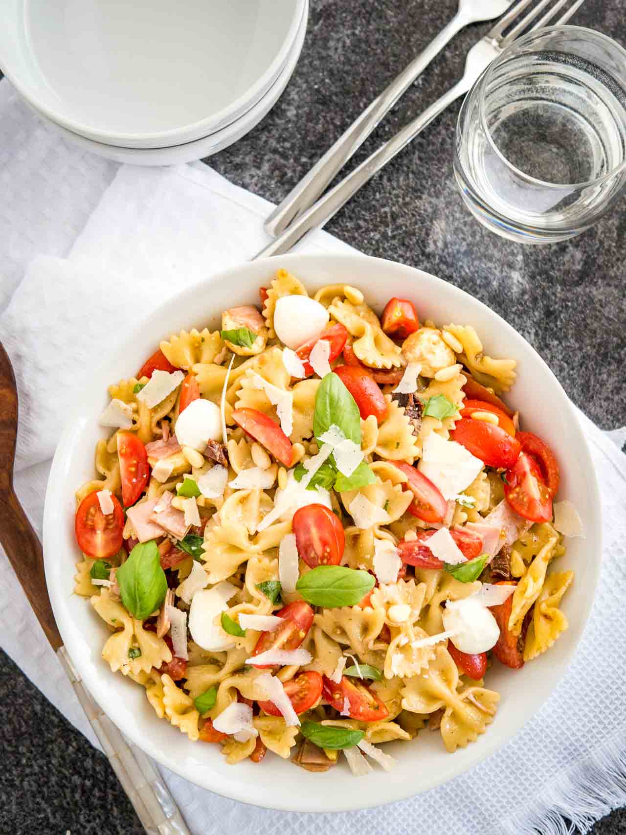 Vue du dessus d'un bol blanc avec une salade de pâtes avec vinaigrette italienne, avec des tomates, de la mozzarella, des feuilles de basilic et des copeaux de parmesan. Le bol est posé sur un torchon blanc avec des fourchettes, un verre d'eau et une pince à salade à côté.