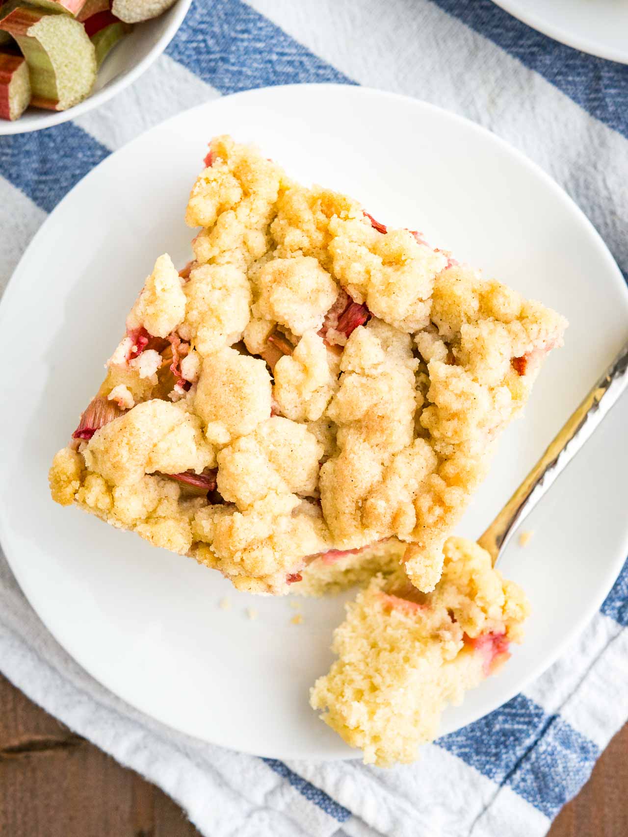 Vue de haut d'un morceau de gâteau au café à la rhubarbe surmonté de streusel assis sur une assiette blanche posée sur un torchon blanc et bleu. Une fourchette a pris une part du gâteau.