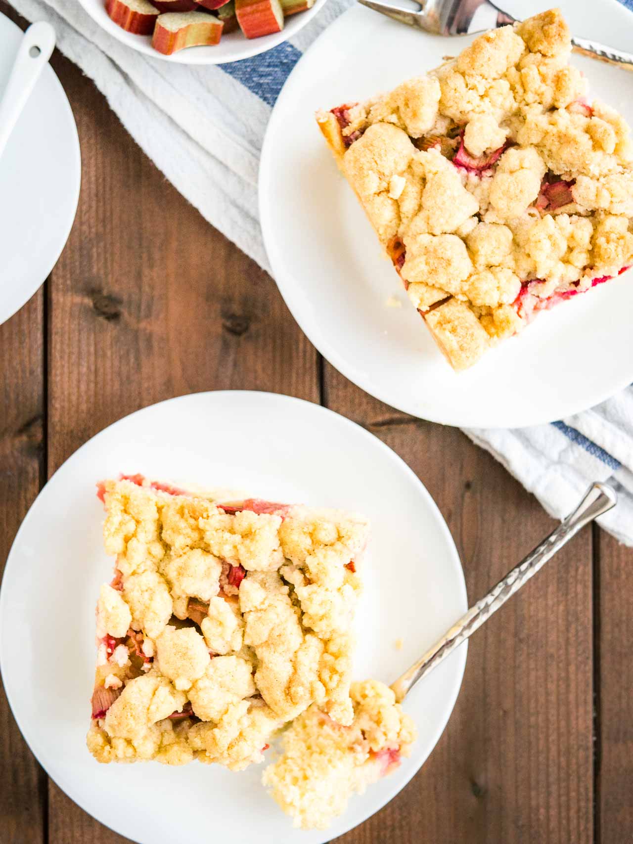 Vue de haut d'une part de gâteau au café à la rhubarbe surmontée de streusel sur une assiette blanche. Une fourchette a pris une part du gâteau. À côté, une autre part de gâteau sur une assiette, un petit bol de rhubarbe et une tasse de café.