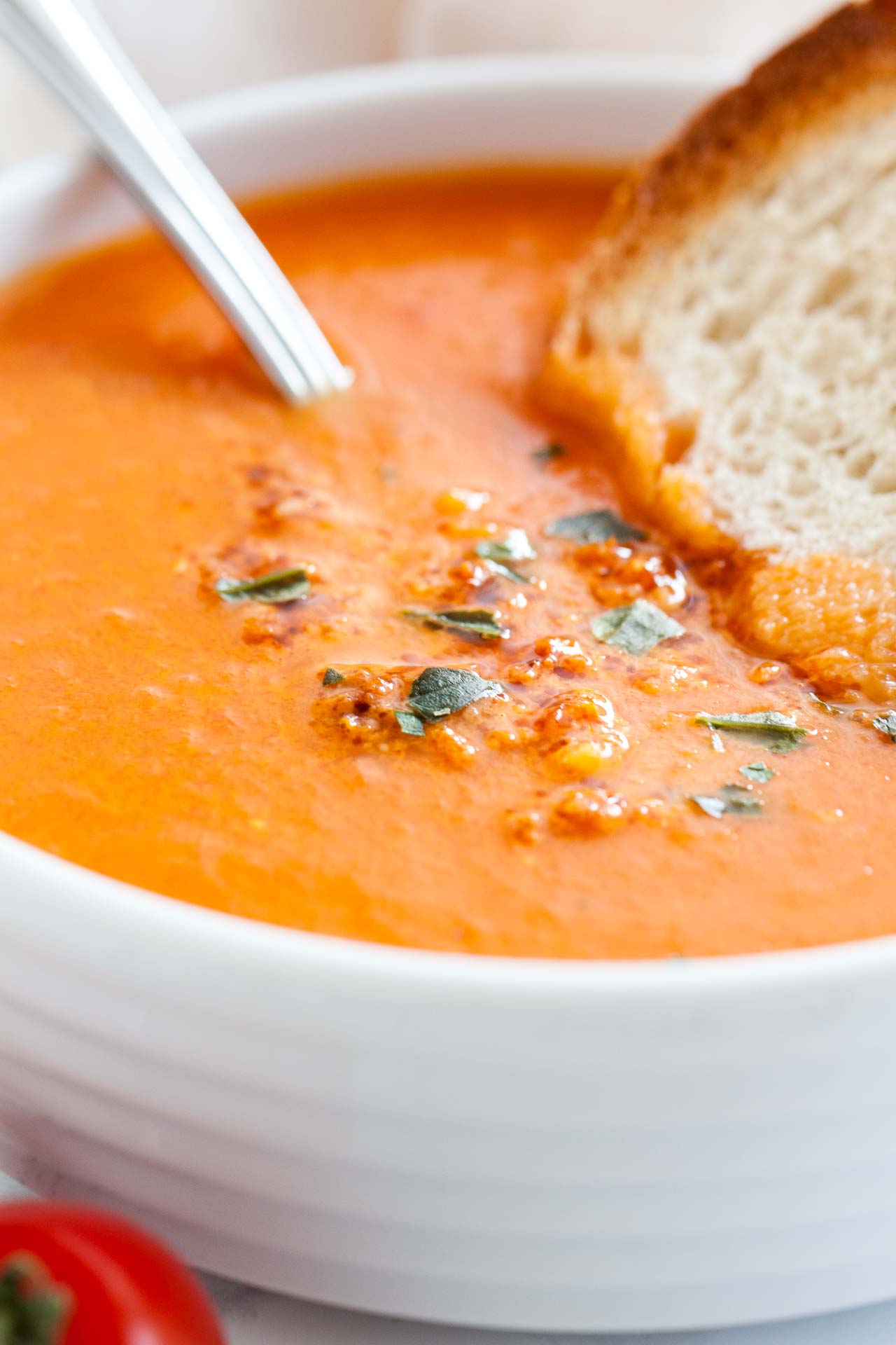 Gros plan d'un bol blanc de soupe de tomates rôties garni de basilic, avec une cuillère et une tranche de pain grillé.