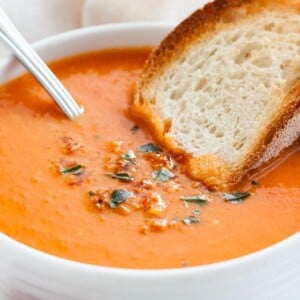 Un bol blanc de soupe de tomates rôties garni de basilic, avec une cuillère et une tranche de pain grillé sur un torchon blanc.