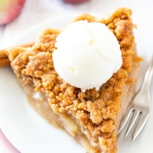 Une part de tarte aux pommes avec un streusel sur une assiette blanche empilée sur une assiette rose avec une fourchette, garnie de glace à la vanille, décorée de bâtons de cannelle et de pommes.