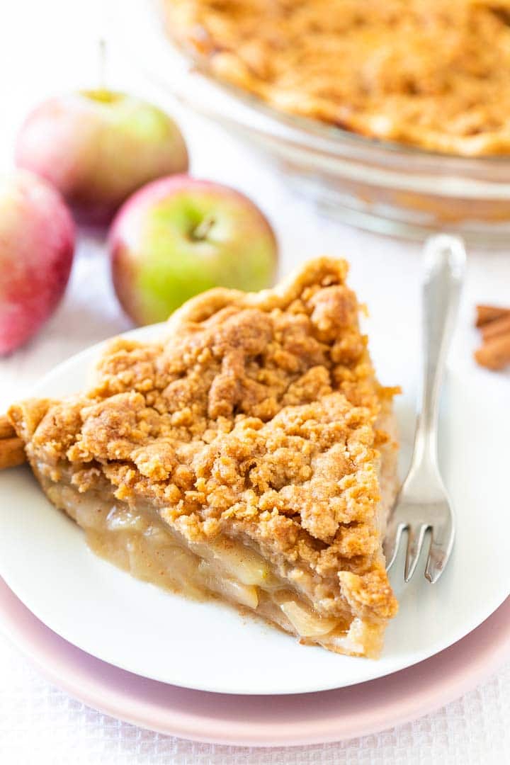 Une part de tarte aux pommes avec un streusel sur une assiette blanche empilée sur une assiette rose avec une fourchette, garnie de glace à la vanille, décorée de bâtons de cannelle, de pommes et un plat en verre en arrière-plan.