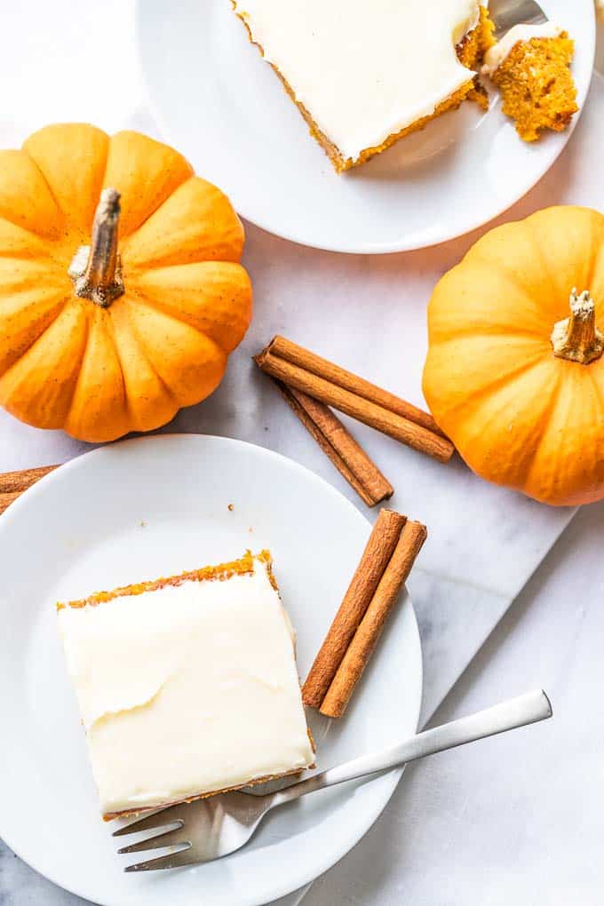 Vue de haut d'un morceau de gâteau à la citrouille recouvert de glaçage et d'une noix de pécan sur une assiette blanche. Une fourchette a pris un morceau et est posée à côté. Il y a 4 morceaux de cannelle et deux petites citrouilles à côté.