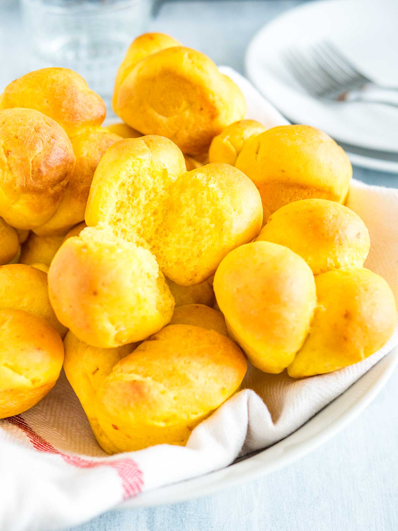 Des petits pains à la citrouille sur un torchon blanc avec une bande rouge sur une assiette blanche. L'un des petits pains a un morceau manquant et il y a une pile d'assiettes blanches avec des fourchettes en arrière-plan.