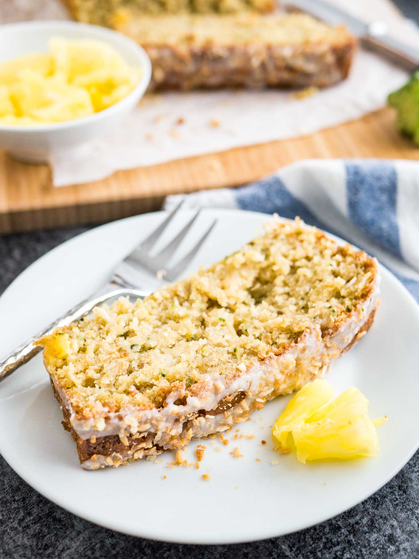 Une assiette blanche avec une tranche de pain à la courgette et à l'ananas avec un peu d'ananas et une fourchette. Le reste du pain est en arrière-plan sur une planche à découper en bois.