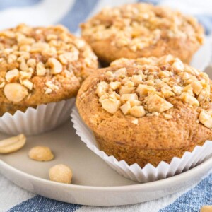 3 muffins à la banane et au beurre de cacahuète, garnis de cacahuètes avec leurs caissettes ouvertes sur une assiette grise sur un torchon blanc et bleu.